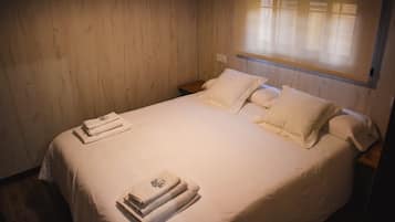 A neatly made bed with white linens and pillows, a wooden headboard, and a window with blinds.