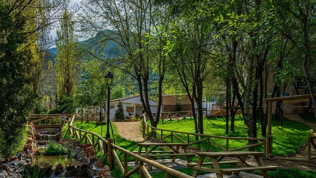 A rustic wooden bridge over a small stream, picnic tables, and a stone building in a lush green park.