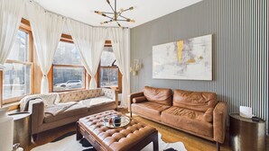 Uma sala de estar moderna com um sofá de couro marrom, um banquinho estofado, uma mesa de centro e um grande quadro abstrato na parede.