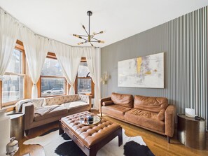 Ein modernes Wohnzimmer mit einem braunen Ledersofa, einem gepolsterten Hocker, einem Couchtisch und einem großen abstrakten Gemälde an der Wand.