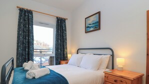 A neatly made bed with white and blue bedding, a wooden nightstand with a lamp, and a window with curtains. - Brewery View (Weymouth)
