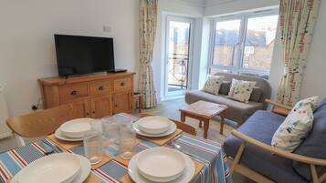 A living room with a dining table set for four, a sofa, a television, and a window with curtains.