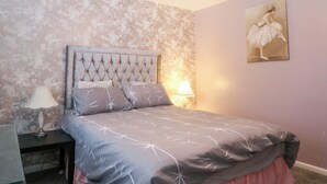 Un dormitorio con cabecera capitonada, cama con colcha estampada, lámpara de noche y un cuadro en la pared.