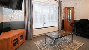 Una sala de estar con televisor, un mueble de madera, un sofá negro, una mesa de centro de vidrio y una araña.