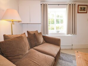 A cozy room with a brown sofa, a lamp, a window with curtains, and a radiator. - Hop Cottage (Petersfield)