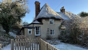 A traditional thatched-roof house with a wooden fence in front, surrounded by snow-covered trees and a clear sky. - Hop Cottage (Petersfield)