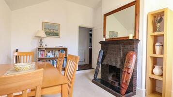 A dining area with a wooden table and chairs, a bookshelf, a fireplace with a mirror above it, and a small shelf with decorative items.