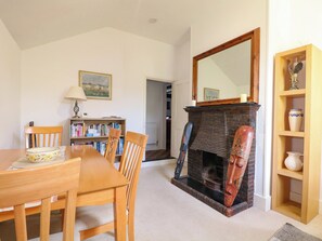 A dining area with a wooden table and chairs, a bookshelf, a fireplace with a mirror above it, and a small shelf with decorative items. - Hop Cottage (Petersfield)