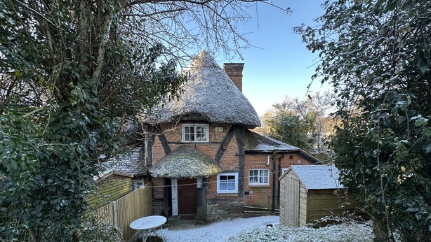 A traditional thatched-roof house with a brick exterior, a wooden door, and a small round table in the front yard. - Hop Cottage (Petersfield)