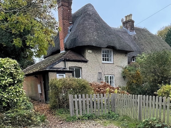 A traditional thatched-roof house with a chimney, surrounded by greenery and a wooden fence.