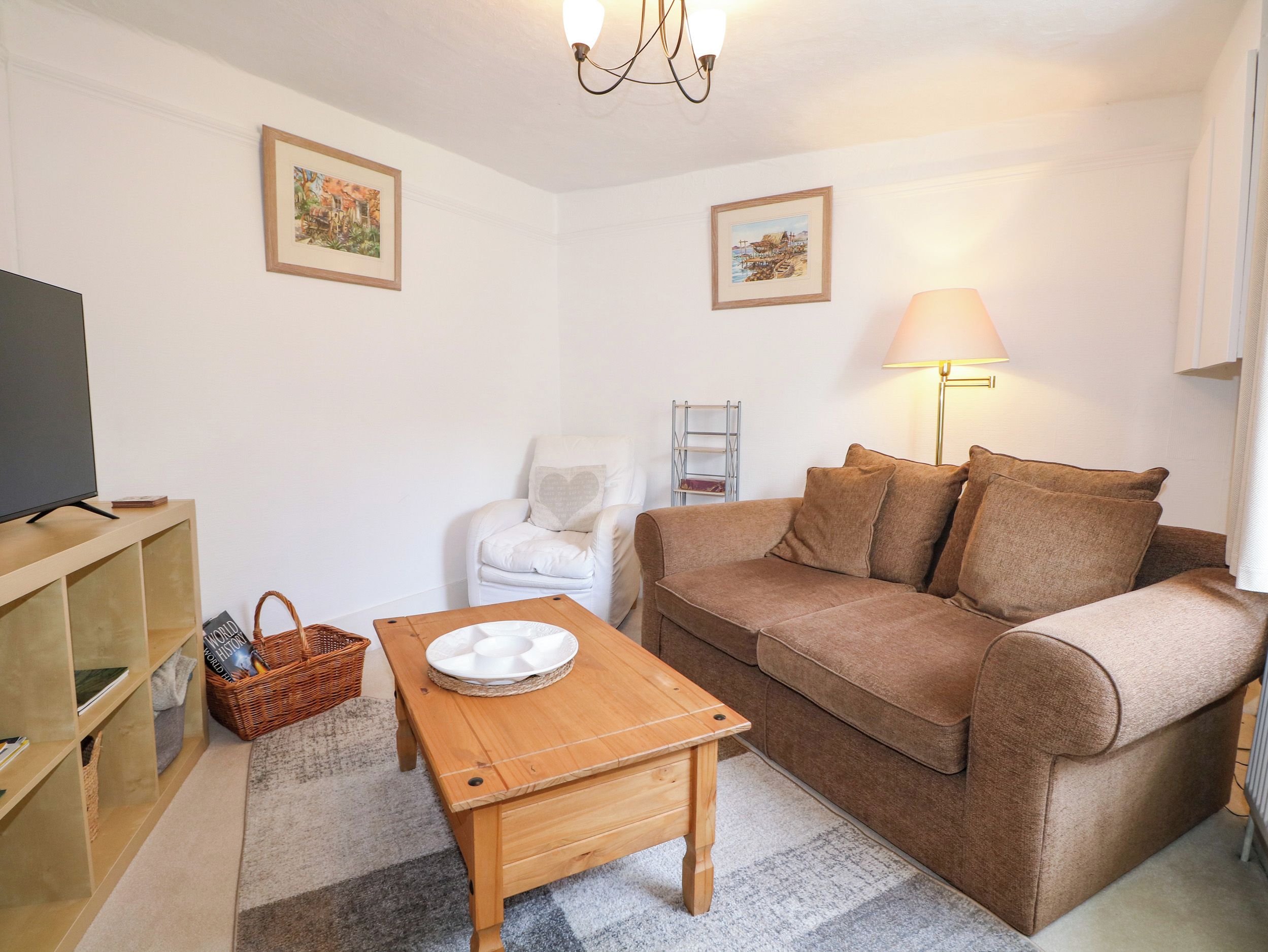 Un salon comprenant un canapé, une table basse en bois, un téléviseur, un fauteuil blanc, une lampe et deux tableaux encadrés accrochés au mur.