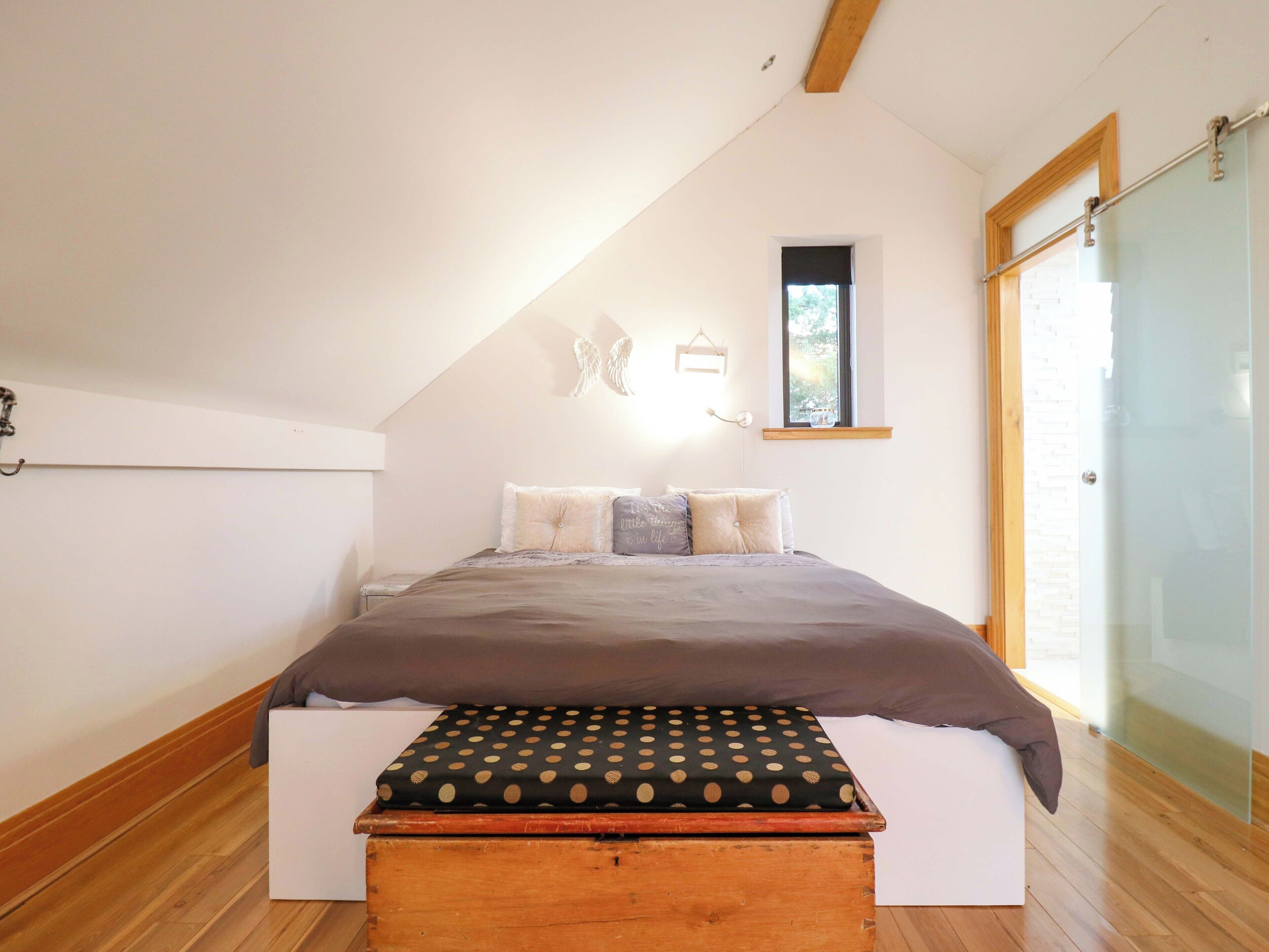 A bedroom with a bed, a window, a wooden bench, and a wall-mounted shelf.