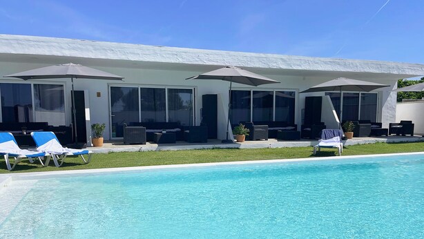 Un área junto a la piscina con sillas de descanso, sombrillas y un edificio con ventanas amplias y un porche cubierto.