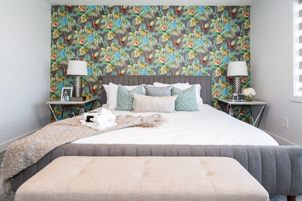 A bedroom with a floral wallpaper, a grey upholstered headboard, and a bedside table with a lamp and decorative items.