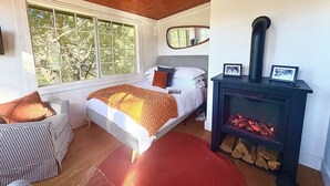 A cozy bedroom with a large window, a bed with a checkered headboard, a red rug, and a wood-burning stove.