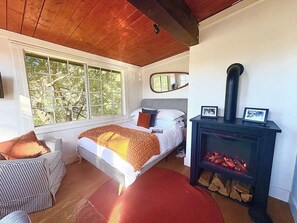 A cozy bedroom with a large window, a bed with a checkered headboard, a red rug, and a wood-burning stove.