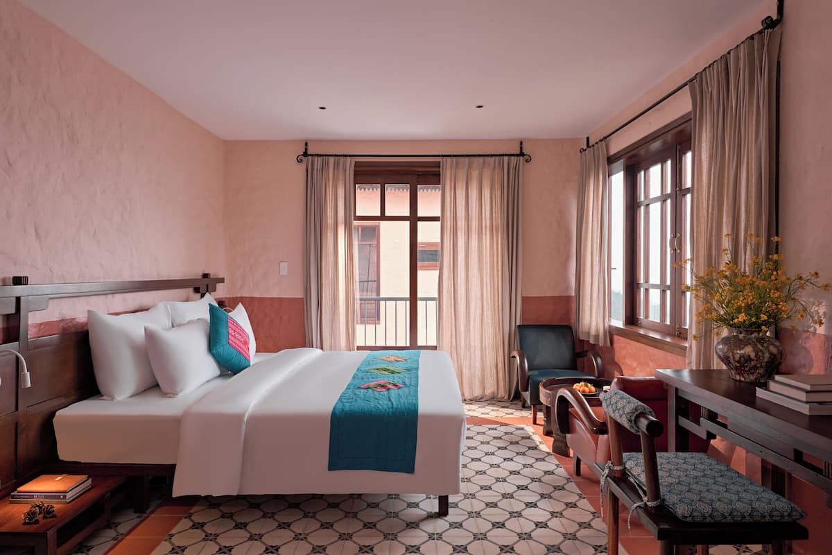 A hotel room with a bed, a chair, a desk, a window with curtains, and a vase with flowers.