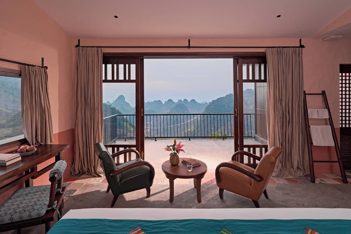 A room with a bed, two chairs, a small table with a vase, and a balcony with a view of mountains.