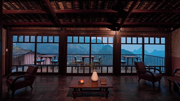 A room with a wooden ceiling, large windows with a mountain view, a table with a vase and book, and two chairs. - Ha Giang Aya Lodge (Lung Phin)