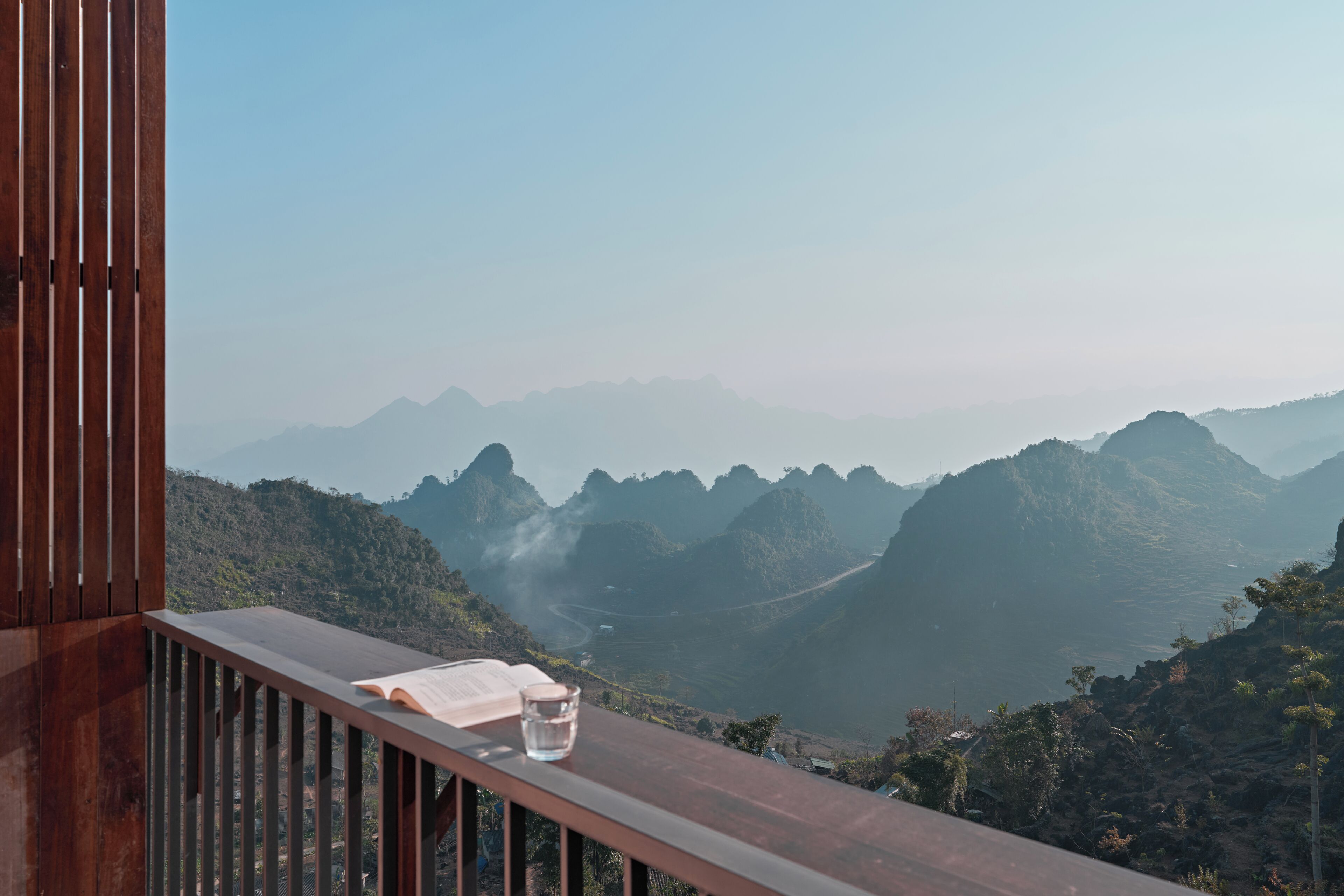 Un balcon en bois avec un livre et un verre d’eau, donnant sur un paysage montagneux.