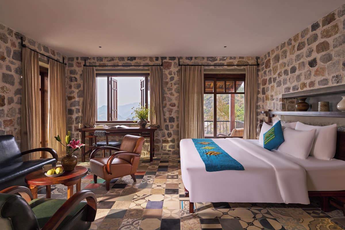 A hotel room with a large bed, a desk, two chairs, and a view of mountains through the window.