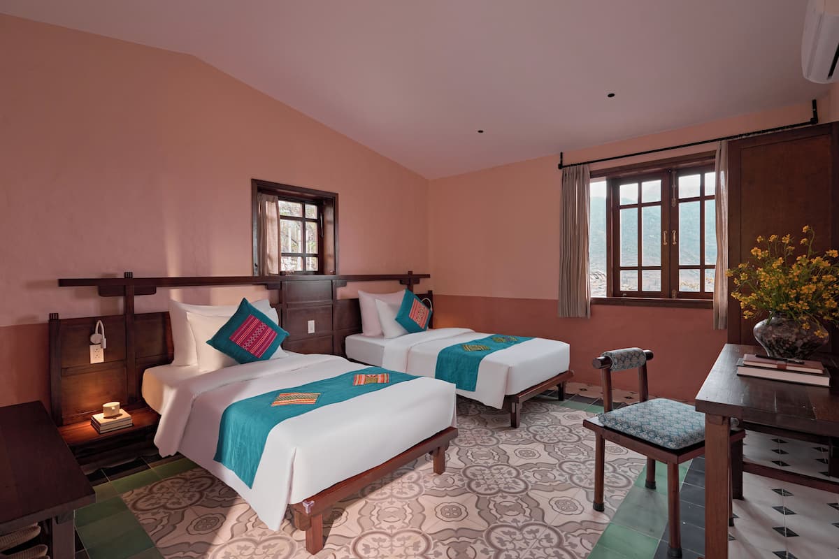 A hotel room with two beds, a desk, a chair, a window with curtains, and a view of the mountains.