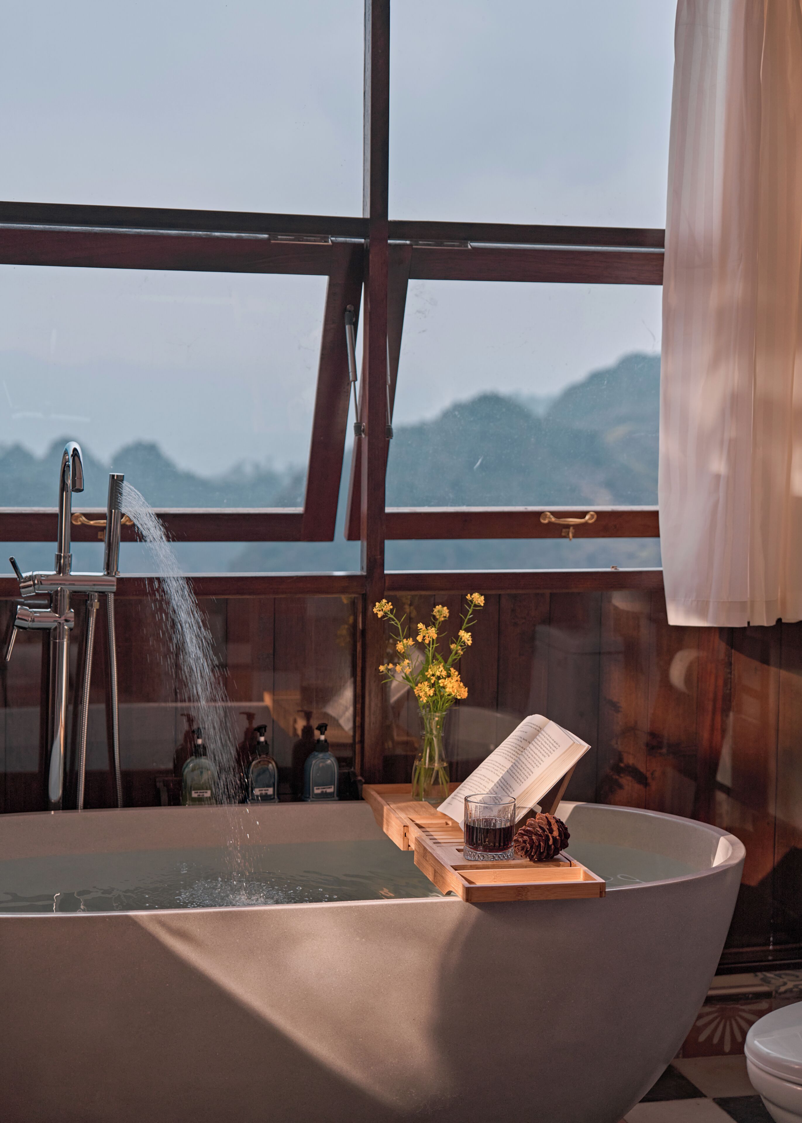 Una bañera con vistas a montañas, una ventana con cortina y un libro sobre una bandeja de madera.