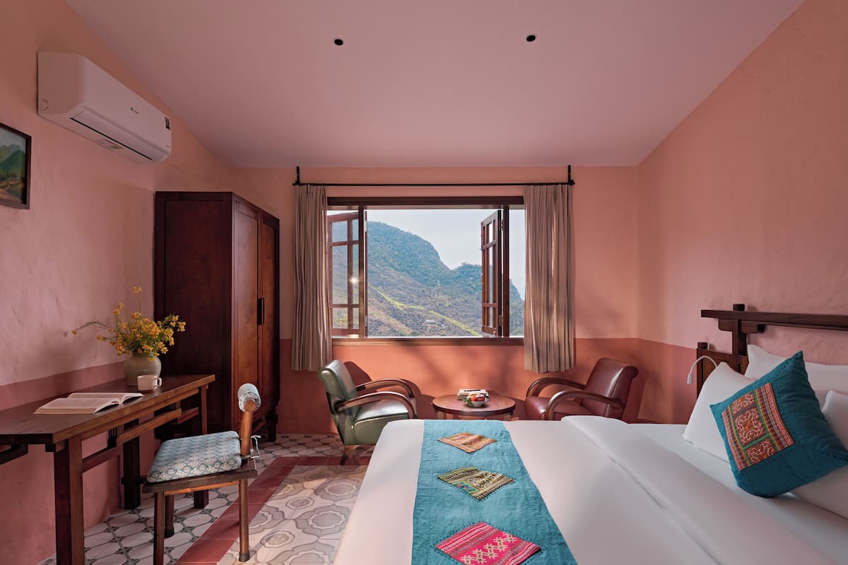 A hotel room with a bed, a desk, a chair, a small table, and a window with a mountain view.