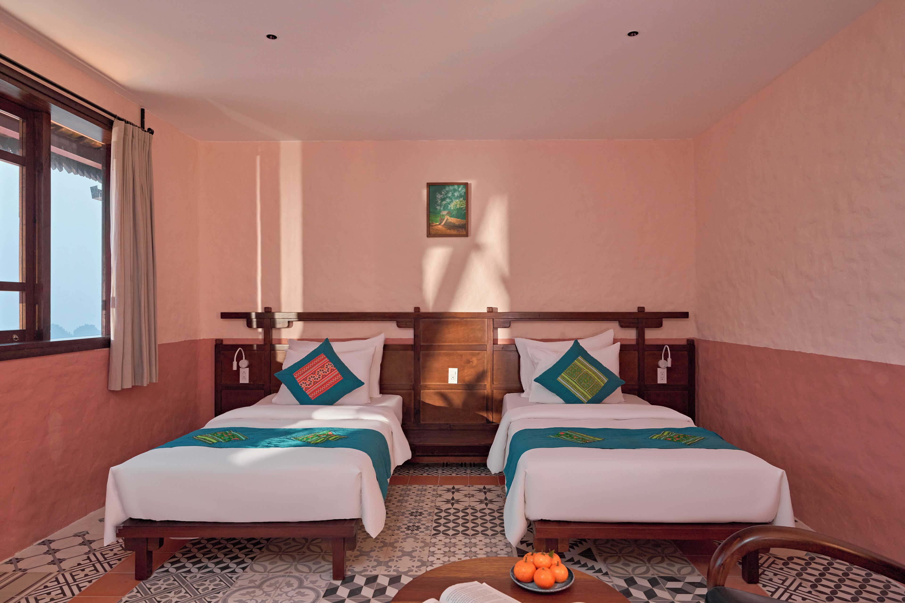 A hotel room with two beds, a small table with oranges, and a painting on the wall.