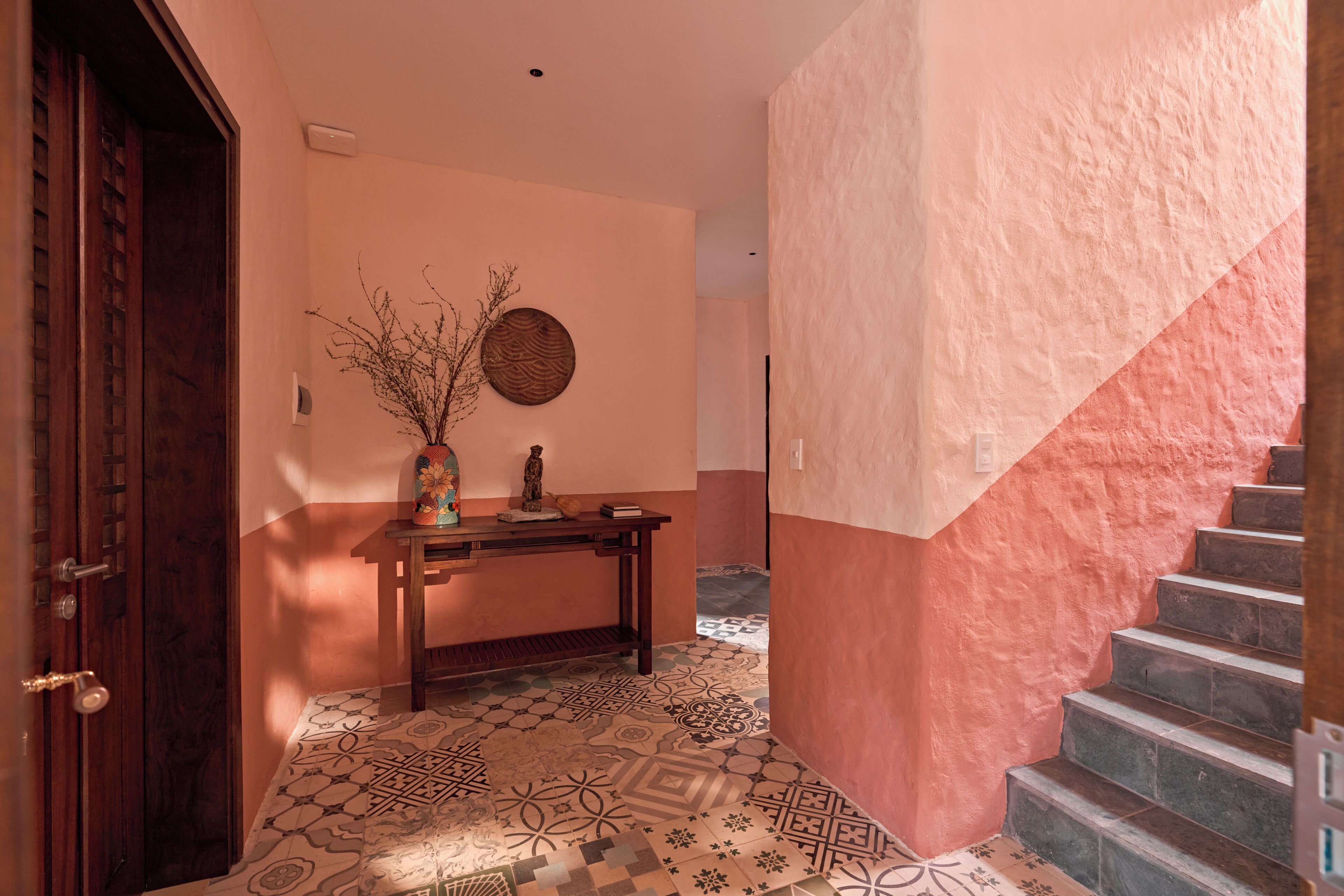 A hallway with patterned floor tiles, a wooden door, a small table with a vase, and a staircase with a pink wall.