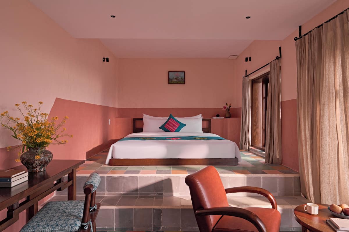 A hotel room with a bed, a chair, a table, a vase with flowers, and a window with curtains.