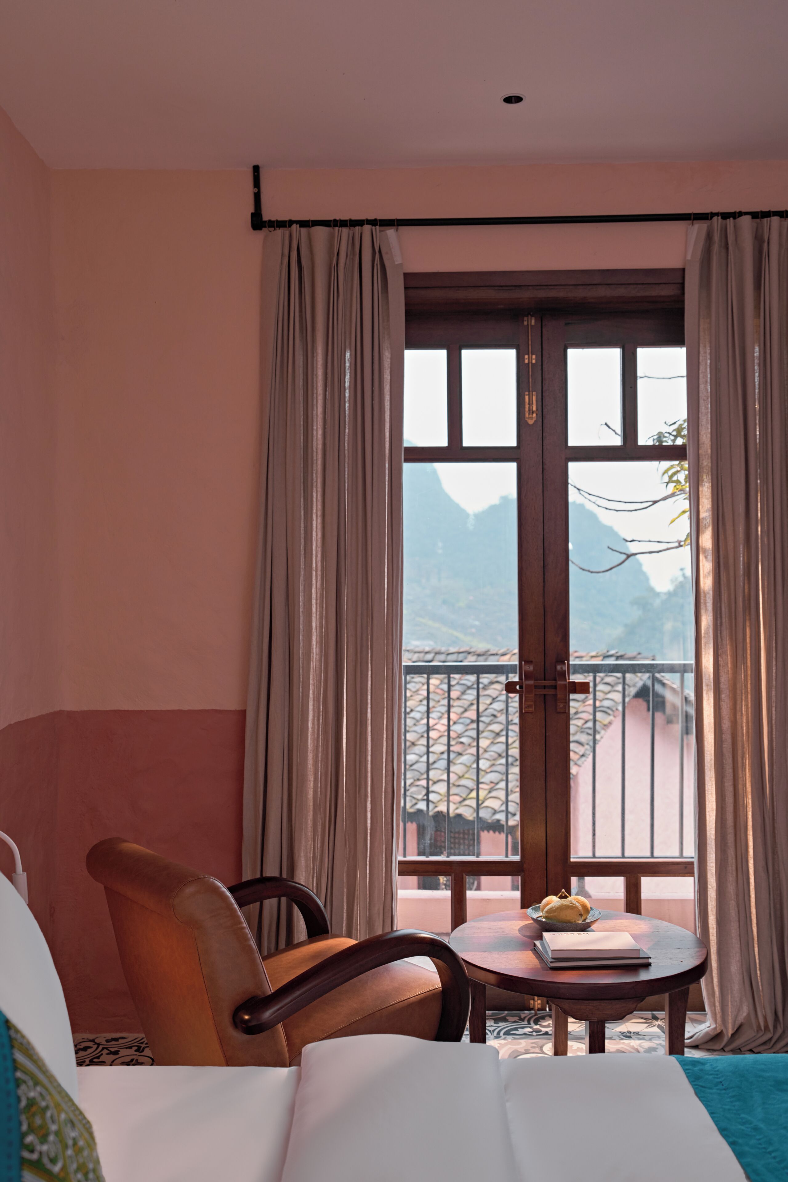 A hotel room with a bed, a chair, a small table with a book and a bowl, and a window with curtains.