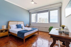 A bedroom with a wooden bed, a nightstand, a desk, and a window with a city view. - Beira-Mar com 5 Quartos e vista incrível (Guarujá)