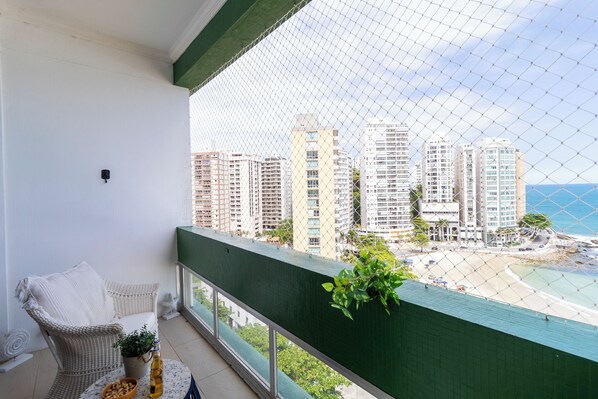 A balcony with a wicker chair, a small table with a plant and a bottle, and a view of high-rise buildings and a beach. - Beira-Mar com 5 Quartos e vista incrível (Guarujá)