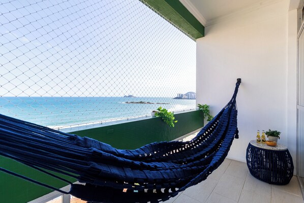 A hammock on a balcony with a view of the sea, a small round table with bottles, and a potted plant. - Beira-Mar com 5 Quartos e vista incrível (Guarujá)