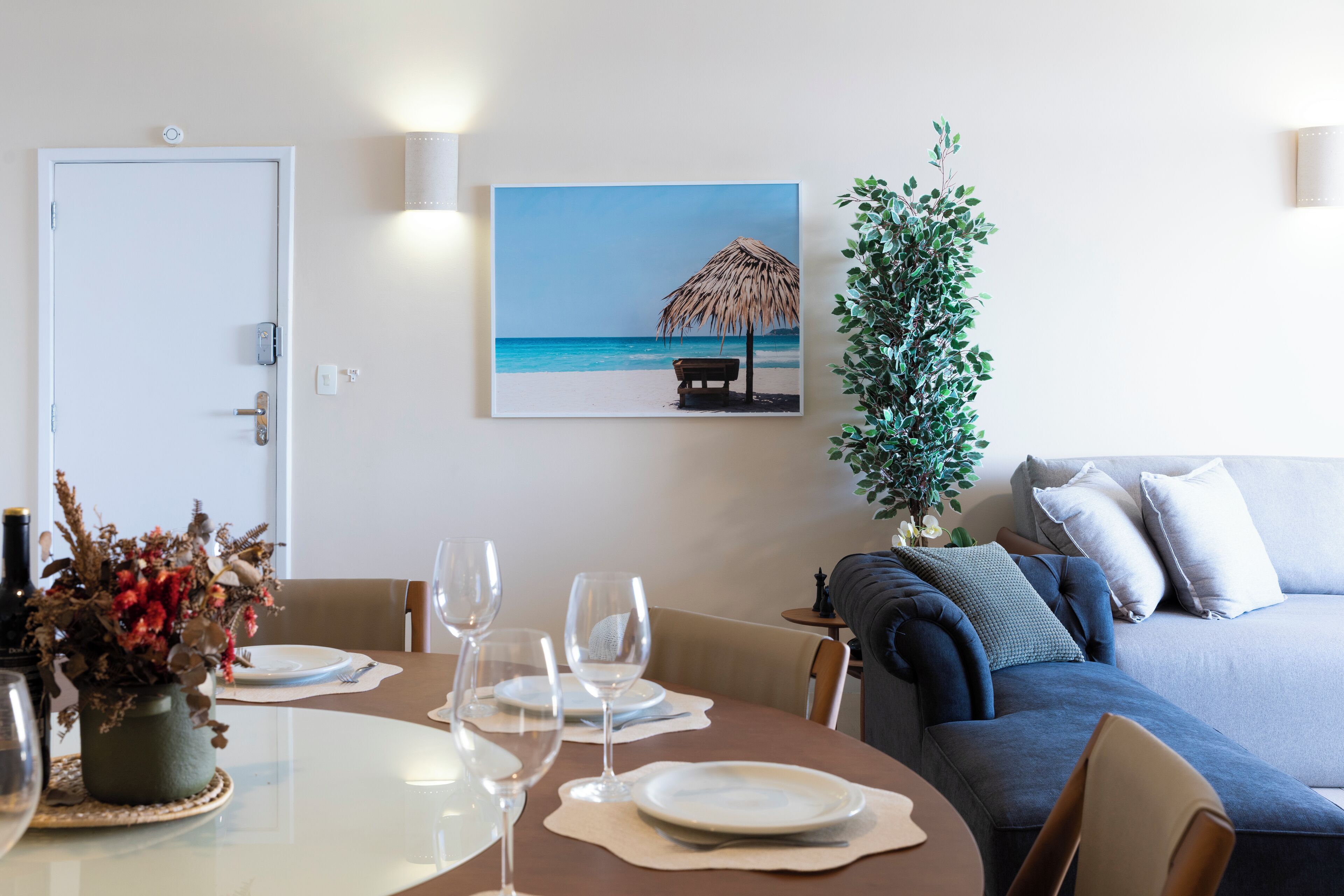 A dining area with a table set for four, a sofa, a potted plant, and a beach-themed painting on the wall.