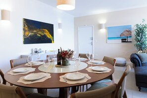 A dining area with a round table set for six, surrounded by chairs. There are two paintings on the walls, a potted plant, and a sofa in the background. - Beira-Mar com 5 Quartos e vista incrível (Guarujá)