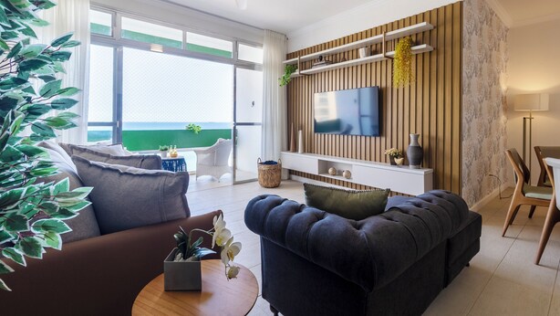 A modern living room with a sofa, a coffee table, a TV mounted on a wooden wall, and a balcony with a view. - Beira-Mar com 5 Quartos e vista incrível (Guarujá)