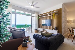 A modern living room with a sofa, a coffee table, a TV mounted on a wooden wall, and a balcony with a view. - Beira-Mar com 5 Quartos e vista incrível (Guarujá)