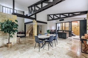 Un vaste espace de vie doté d’un haut plafond, de poutres apparentes, d’une table à manger avec des chaises, d’un escalier et d’un coin cuisine.