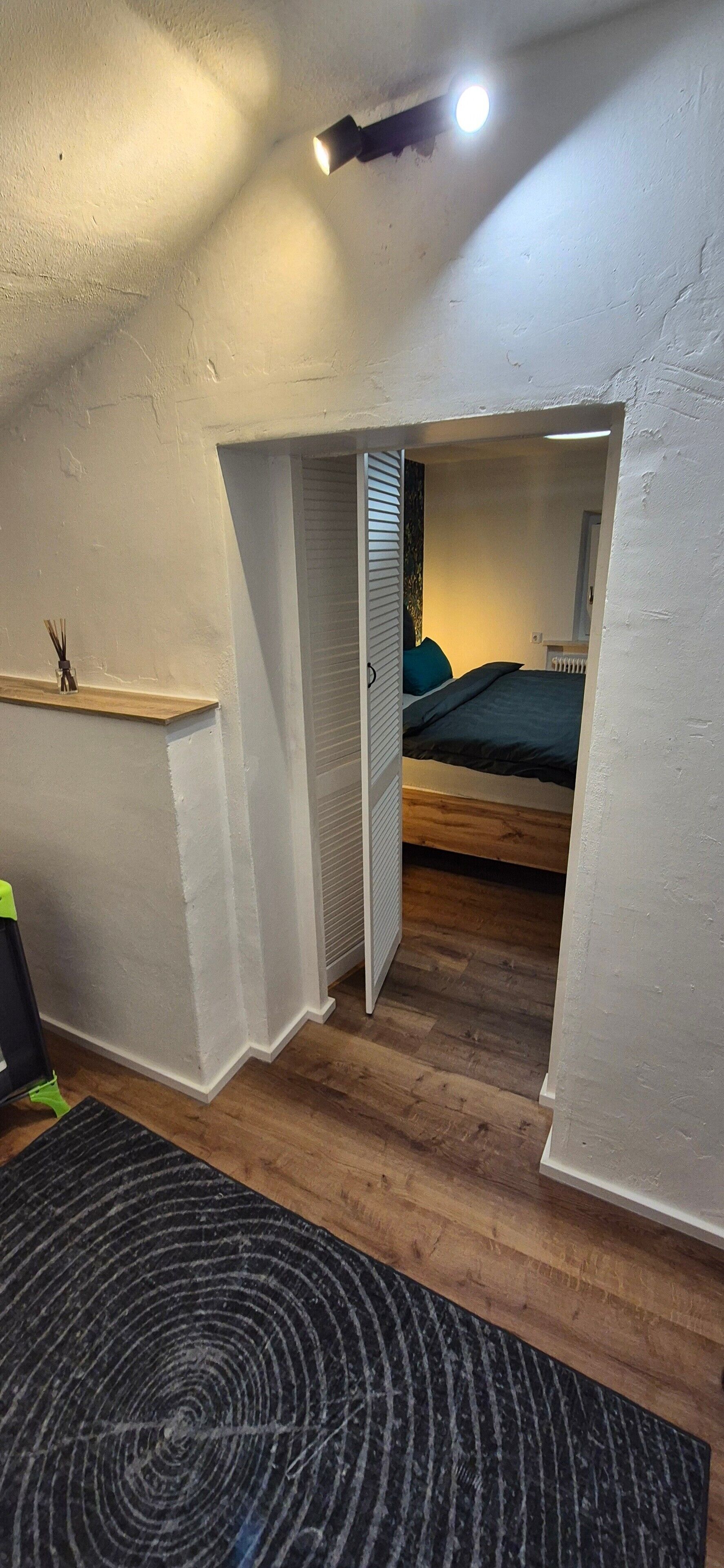 A room with a sloped ceiling, a bed with a green blanket, a small shelf with a bottle, and a black circular rug.