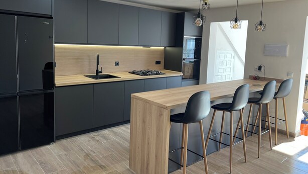 A modern kitchen with dark cabinetry, a wooden countertop, and a built-in stove. There are three bar stools and a large window providing natural light. - Villa Palma Cabo Roig- Spacious 5-bedroom villa in peaceful Cabo Roig with AC (orihueala costa)