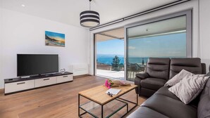 Una sala de estar moderna con un televisor de pantalla plana, un sofá, una mesa de centro y un amplio ventanal corredera que da a un balcón con vistas al mar.
