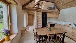 A modern loft-style room with a wooden ceiling, a dining area with a table and chairs, a kitchen with white countertops, and a view of the outdoors through a window. - Stone house near the ocean (Tréogat)
