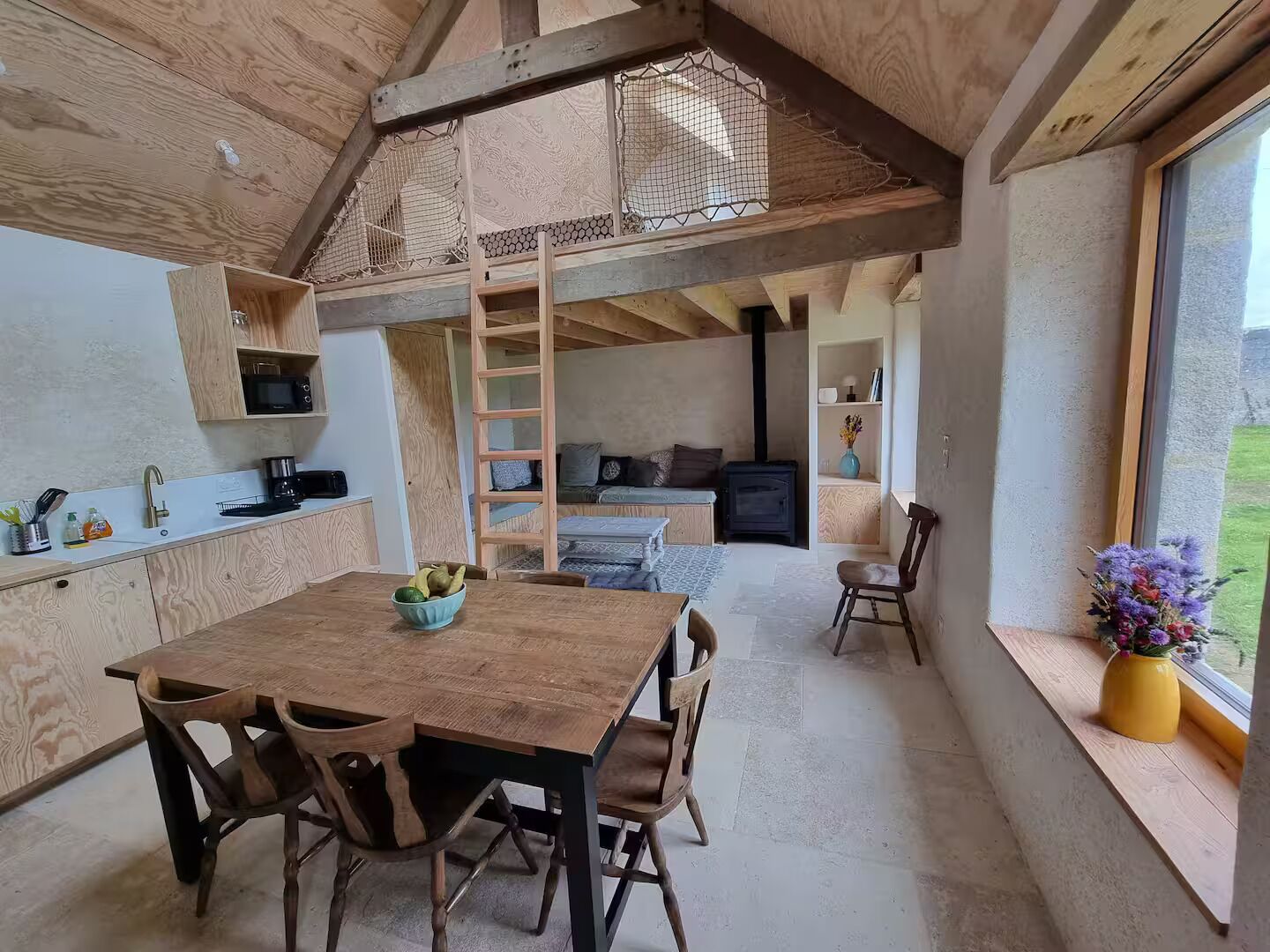 Ein rustikales Holzinnere mit Esstisch, Stühlen, einer Küchenzeile, einem Schlafplatz in der Mansarde und einem großen Fenster mit Blick ins Freie.