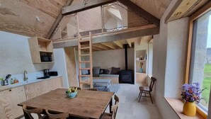 A rustic wooden interior with a dining table, chairs, a kitchen area, a loft bed, and a large window with a view of the outdoors. - Stone house near the ocean (Tréogat)
