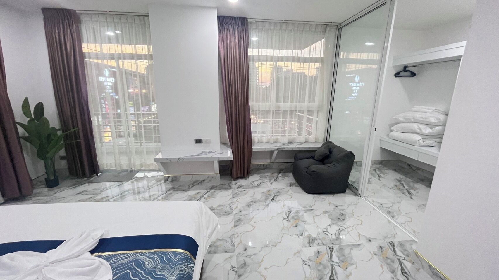A modern hotel room with a marble floor, a bed, a seating area, a bathtub, and a view of the city through large windows.