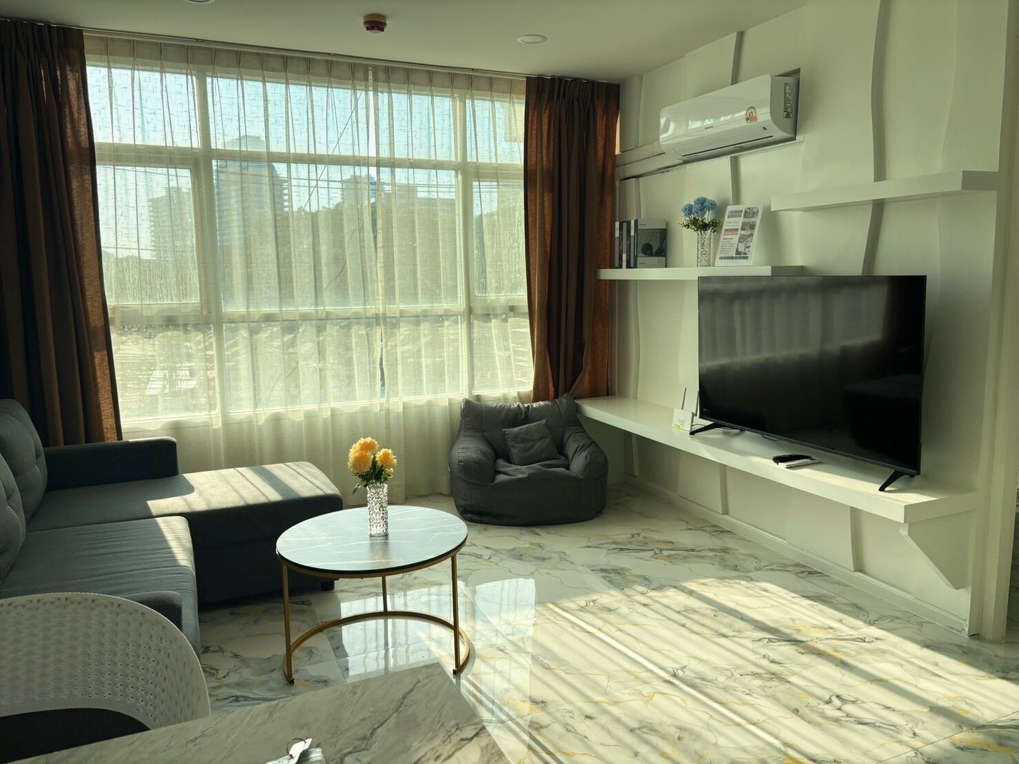A modern living room with a sofa, a round coffee table, a grey armchair, a flat-screen TV, and a large window with sheer curtains.