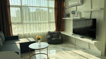 A modern living room with a sofa, a round coffee table, a grey armchair, a flat-screen TV, and a large window with sheer curtains.