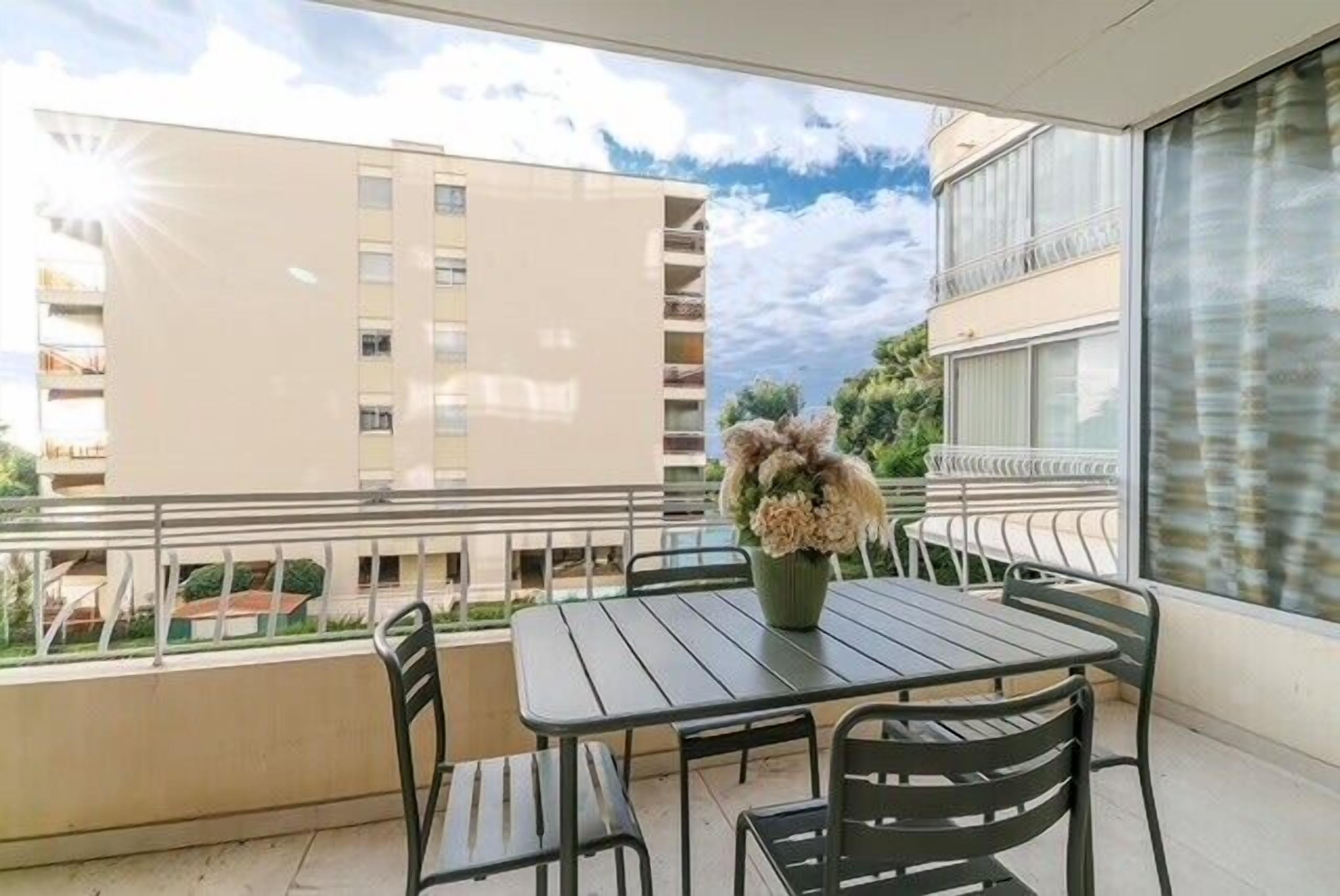 A balcony with a table and chairs, a vase with flowers, and a view of a building and greenery.
